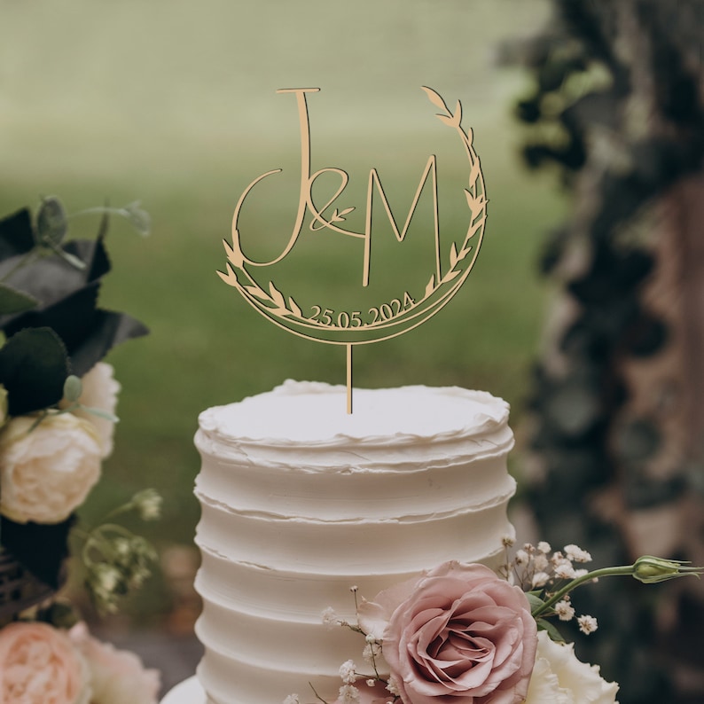 May include: A white wedding cake with a gold wooden topper featuring the letters "J&M" and the date "25.05.2024" surrounded by a wreath of leaves.