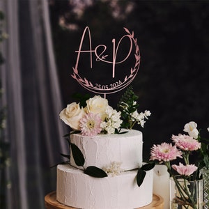 May include: A two-tiered white wedding cake with a rose gold cake topper featuring the letters "A&P" in a script font with a leafy wreath and the date "25.05.2024". The cake is decorated with white and pink flowers.