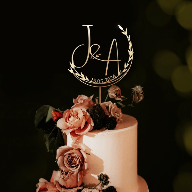 May include: Gold wedding cake topper with the initials "J & A" in a script font surrounded by a wreath of leaves. The date "25.05.2024" is written below the initials.