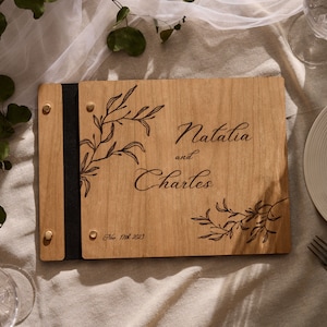May include: A wooden guest book with a black leather strap and brass accents. The cover is engraved with a floral design and the text "Natalia and Charles" and "Oct. 17th 2023".