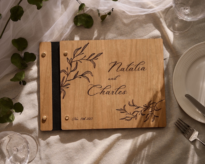 May include: A wooden guest book with a black leather strap and brass accents. The cover is engraved with a floral design and the text "Natalia and Charles" and "Oct. 17th 2023".