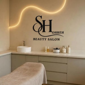 Könnte beinhalten: Ein schwarzes Wand-Schild mit den stilisierten Buchstaben SH, dem Text OHREH und den Worten BEAUTY SALON. Das Schild befindet sich über einer weißen Arbeitsplatte mit Waschbecken, verschiedenen Flaschen und einer beige abgedeckten Liege.