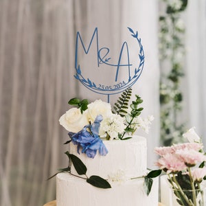 May include: Blue wedding cake topper with the initials M&A and the date 25.05.2024, surrounded by a leafy wreath. The topper is on a white cake with white and blue flowers.