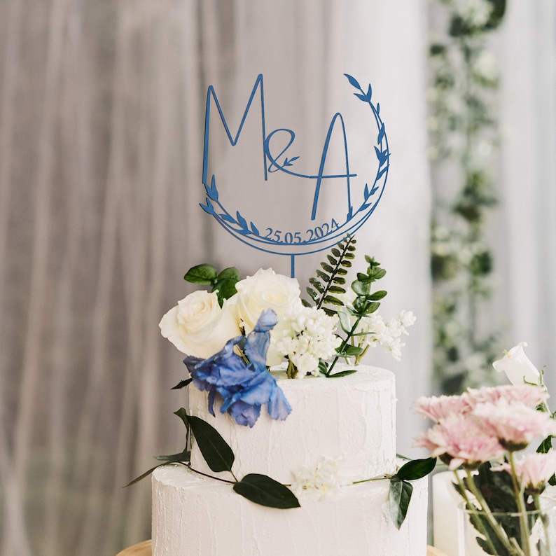 May include: Blue wedding cake topper with the initials M&A and the date 25.05.2024, surrounded by a leafy wreath. The topper is on a white cake with white and blue flowers.