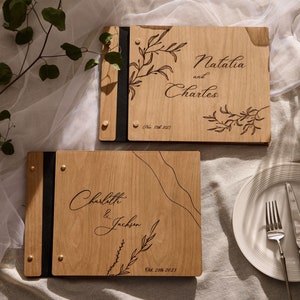 May include: Two wooden guest books with black leather covers and engraved floral designs. The top book says "Natalia and Charles" with the date "Feb 17th 2023". The bottom book says "Charlotte & Jackson" with the date "Oct 23rd 2023".