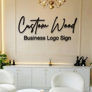 May include: A wall-mounted business logo sign featuring the words Custom Wood in a black cursive script font, with the words Business Logo Sign in a black sans-serif font positioned directly underneath on a light-coloured wall surface.