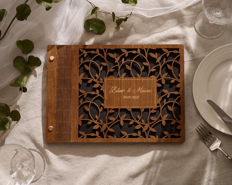 May include: A wooden guest book with a cut-out design of leaves and vines. The cover is personalized with the names "Blair & Mason" and the date "01.01.2023".