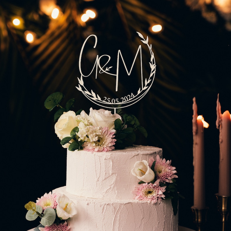 May include: A two-tiered white wedding cake decorated with white and pink flowers. A white wooden cake topper with the letters "C&M" and the date "25.05.2024" is on top of the cake.