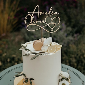 Könnte beinhalten: Ein Kuchenaufsatz aus Holz mit den Namen "Amelia" und "Oliver", einem Herz und dem Datum 10.05.2026. Der Aufsatz befindet sich auf einer weißen, mit Makronen, Blumen und Zitronenscheiben dekorierten Torte.