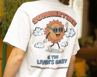 Puede incluir: Camiseta blanca con un gráfico de un sol sonriente con gafas de sol, nubes y el texto "SUMMERTIME & THE LIVIN'S EASY". La camiseta es de manga corta y de corte holgado. La modelo lleva un sombrero blanco.