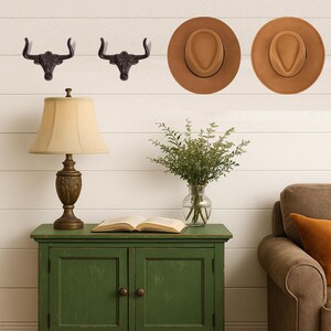 May include: Two dark metal wall hooks shaped like bull heads, two tan felt fedora hats, a table lamp with a beige shade, a glass vase with green foliage, and an open book on a green wooden cabinet with two doors and round knobs.