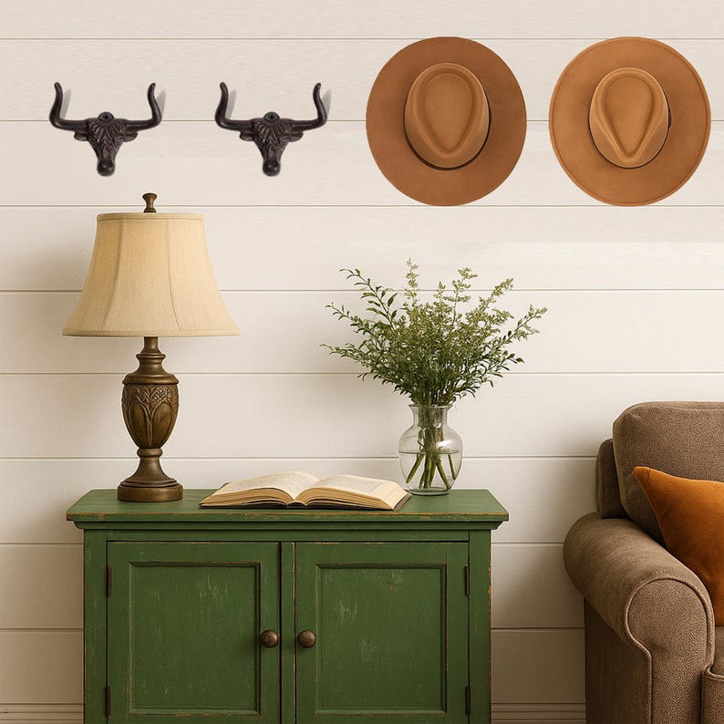 May include: Two dark metal wall hooks shaped like bull heads, two tan felt fedora hats, a table lamp with a beige shade, a glass vase with green foliage, and an open book on a green wooden cabinet with two doors and round knobs.