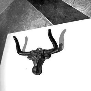 May include: A black cast iron wall hook shaped like a bull head with prominent curved horns. The metal surface features a textured finish and a single mounting hole at the top center, designed for hanging items on a flat vertical surface.