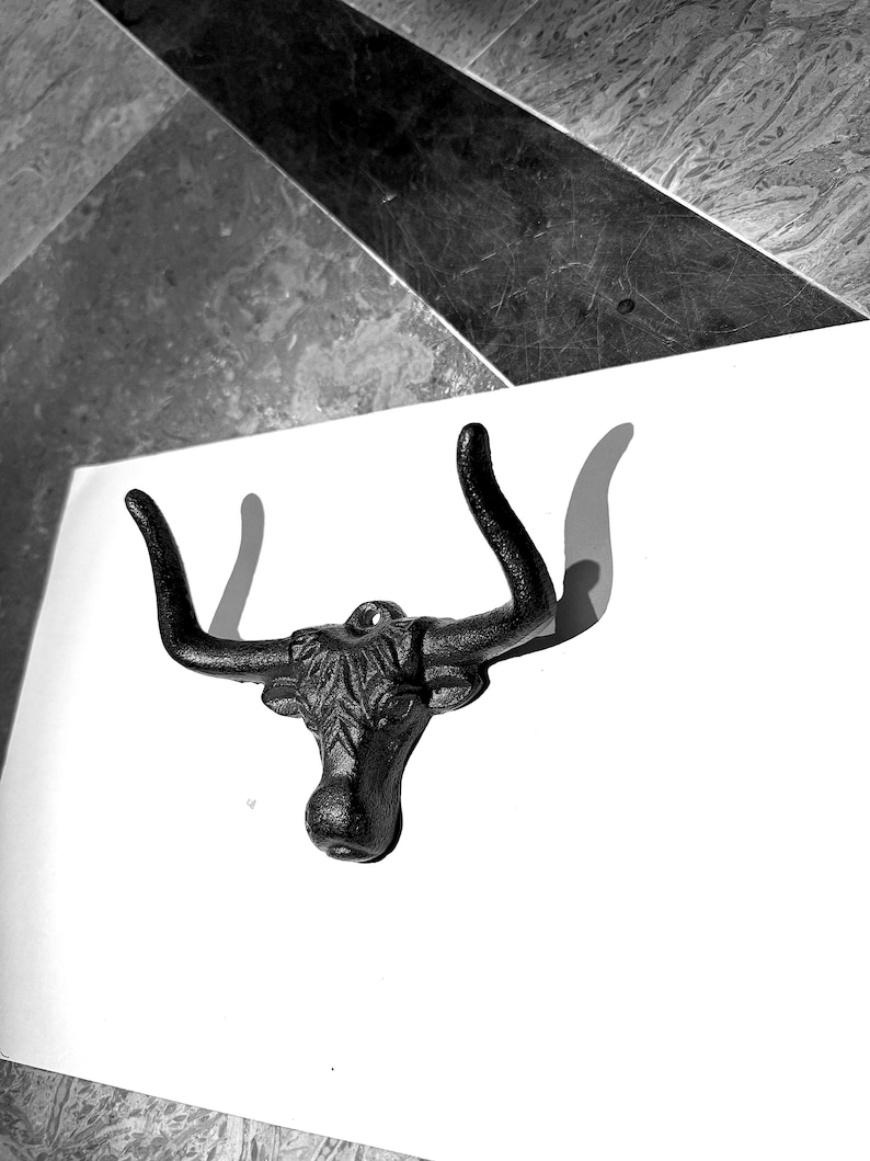 May include: A black cast iron wall hook shaped like a bull head with prominent curved horns. The metal surface features a textured finish and a single mounting hole at the top center, designed for hanging items on a flat vertical surface.