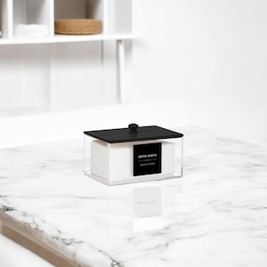 May include: A clear acrylic box with a black lid and a small black knob. The box contains white dryer sheets and has a black label that reads "DRYER SHEETS - LAUNDRY PICKUP". The box is sitting on a white marble surface.