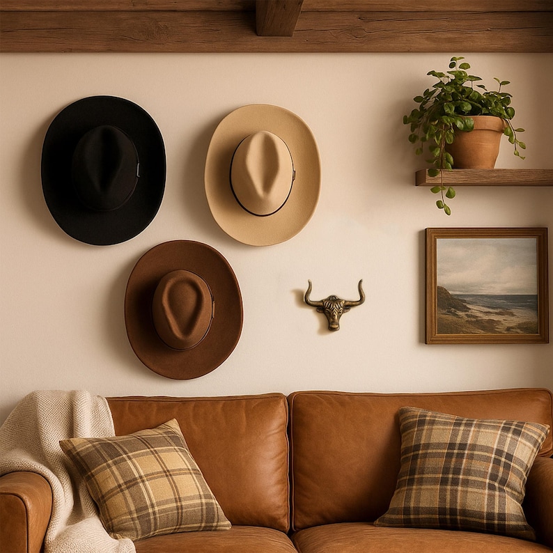 May include: Three felt hats in black, beige, and brown are mounted on a wall above a brown leather sofa with plaid pillows. A brass bull head wall hook, a framed landscape painting, and a potted green plant on a shelf are also visible.