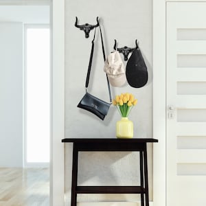 May include: Two black wall-mounted hooks shaped like bull skulls. One hook holds a black crossbody bag, while the other holds a beige baseball cap and a black baseball cap. A yellow vase with yellow tulips sits on a dark wooden table.