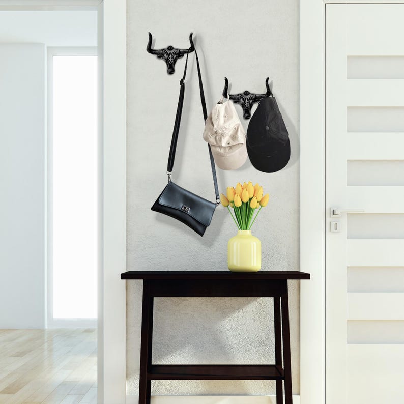 May include: Two black wall-mounted hooks shaped like bull skulls. One hook holds a black crossbody bag, while the other holds a beige baseball cap and a black baseball cap. A yellow vase with yellow tulips sits on a dark wooden table.