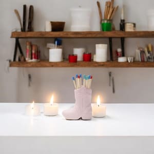 May include: A light pink ceramic cowboy boot-shaped match holder filled with matches with blue and red tips. Three lit candles are in front of the boot. The boot is on a white surface with a wooden shelf in the background.