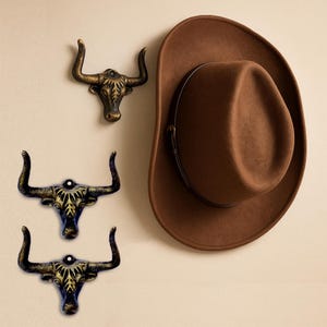 May include: A brown felt hat with a dark brown band and three metallic wall hooks shaped like bull heads. The hooks feature a distressed gold and black finish with prominent horns and a mounting hole at the top of each head.