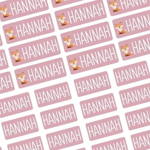 Könnte beinhalten: Rosa rechteckige Etiketten mit dem Namen "HANNAH" in weißen Blockbuchstaben. Jedes Etikett zeigt eine kleine Illustration eines Fuchses. Die Etiketten sind in einem sich wiederholenden Muster angeordnet.