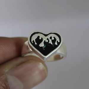 Può includere: Un anello d'argento con un design a forma di cuore nero. Il cuore ha un design bianco di una fiamma stilizzata all'interno.