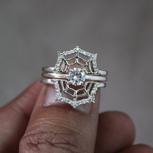 May include: A silver ring with a spiderweb design and a clear gemstone in the center. The ring is set on a finger.