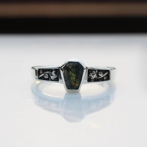 K&ouml;nnte beinhalten: Ein silberner Ring mit einem Sarg-f&ouml;rmigen, dunkelgr&uuml;nen Edelstein in der Mitte. Das Band hat Blumendesigns auf jeder Seite des Edelsteins. Der Ring ist auf einer hellen, reflektierenden Oberfl&auml;che platziert.
