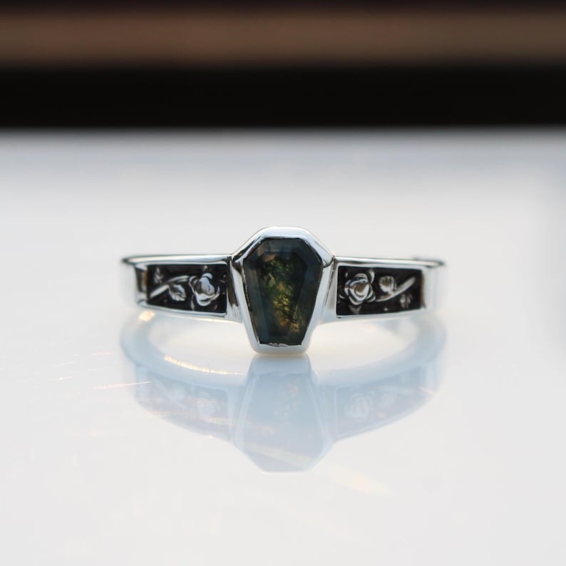 K&ouml;nnte beinhalten: Ein silberner Ring mit einem Sarg-f&ouml;rmigen, dunkelgr&uuml;nen Edelstein in der Mitte. Das Band hat Blumendesigns auf jeder Seite des Edelsteins. Der Ring ist auf einer hellen, reflektierenden Oberfl&auml;che platziert.