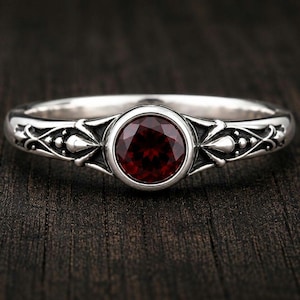 Può includere: Un anello in argento con una pietra preziosa granato rosso scuro, rotonda e sfaccettata, incastonata al centro. La fascia presenta intricati motivi metallici in rilievo che si estendono dall'incastonatura centrale lungo entrambi i lati.