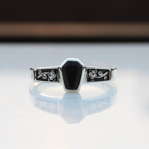 Peut inclure: Bague en argent ornée d'une pierre noire en forme de cercueil. La monture est ornée de motifs de roses et de feuilles de chaque côté du cercueil. La bague est posée sur une surface blanche réfléchissante.