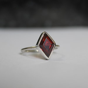 May include: A silver ring with a red gemstone set in a diamond-shaped setting.