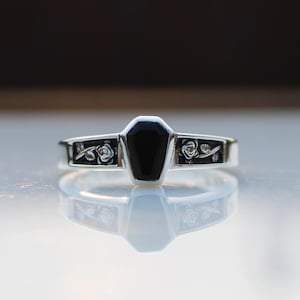 K&ouml;nnte beinhalten: Ein silberner Ring mit einem schwarzen, sargf&ouml;rmigen Stein. Das Band hat florale Details auf beiden Seiten des schwarzen Steins. Der Ring wird auf einer reflektierenden Oberfl&auml;che gezeigt.
