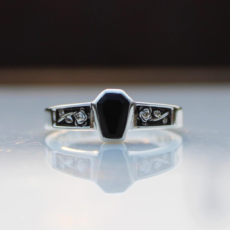 K&ouml;nnte beinhalten: Ein silberner Ring mit einem schwarzen, sargf&ouml;rmigen Stein. Das Band hat florale Details auf beiden Seiten des schwarzen Steins. Der Ring wird auf einer reflektierenden Oberfl&auml;che gezeigt.