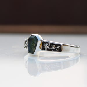 K&ouml;nnte beinhalten: Ein silberner Ring mit einem rechteckigen, gr&uuml;nen Edelstein. Der Ring hat ein Blumenmuster auf dem Band. Der Edelstein hat ein einzigartiges, moosartiges Aussehen. Der Ring wird auf einer reflektierenden Oberfl&auml;che gezeigt.