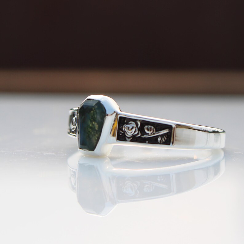 K&ouml;nnte beinhalten: Ein silberner Ring mit einem rechteckigen, gr&uuml;nen Edelstein. Der Ring hat ein Blumenmuster auf dem Band. Der Edelstein hat ein einzigartiges, moosartiges Aussehen. Der Ring wird auf einer reflektierenden Oberfl&auml;che gezeigt.