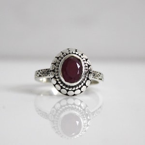 May include: A silver ring featuring an oval ruby gemstone set within an ornate bezel. The ring band is decorated with a delicate scroll pattern.