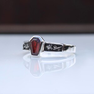 K&ouml;nnte beinhalten: Ein silberner Ring mit einem Sarg-f&ouml;rmigen, tiefroten Edelstein. Das Band ist mit floralen Mustern verziert. Der Ring hat ein gotisches Design und eine polierte Oberfl&auml;che.