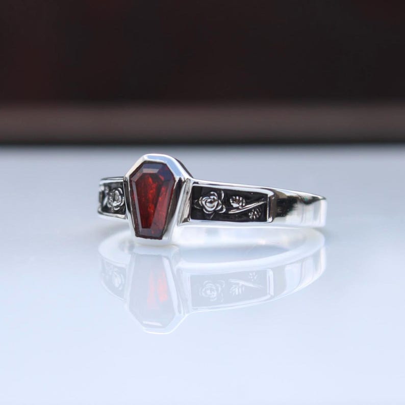 K&ouml;nnte beinhalten: Ein silberner Ring mit einem Sarg-f&ouml;rmigen, tiefroten Edelstein. Das Band ist mit floralen Mustern verziert. Der Ring hat ein gotisches Design und eine polierte Oberfl&auml;che.