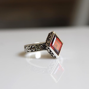 Puede incluir: Un anillo de plata con una gran gema roja tallada en forma de rombo. El anillo tiene una banda grabada y un engaste detallado alrededor de la gema. El anillo se muestra sobre una superficie blanca.