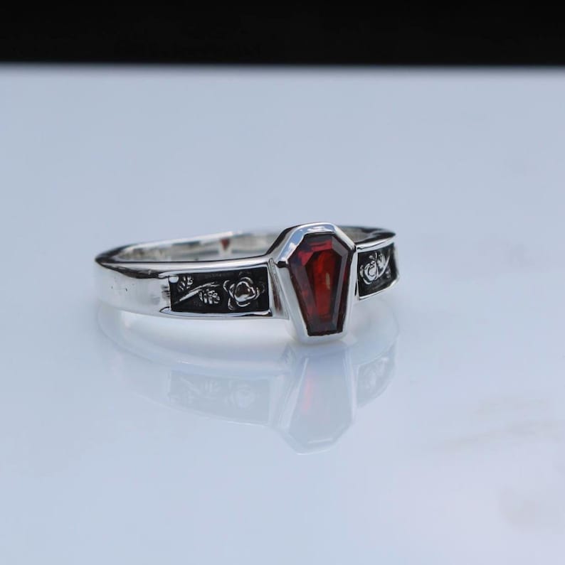 K&ouml;nnte beinhalten: Ein silberner Ring mit einem Sarg-f&ouml;rmigen, tiefroten Edelstein. Das Band ist mit floralen und Rankenmustern verziert. Das Design des Rings ist gotisch und einzigartig.
