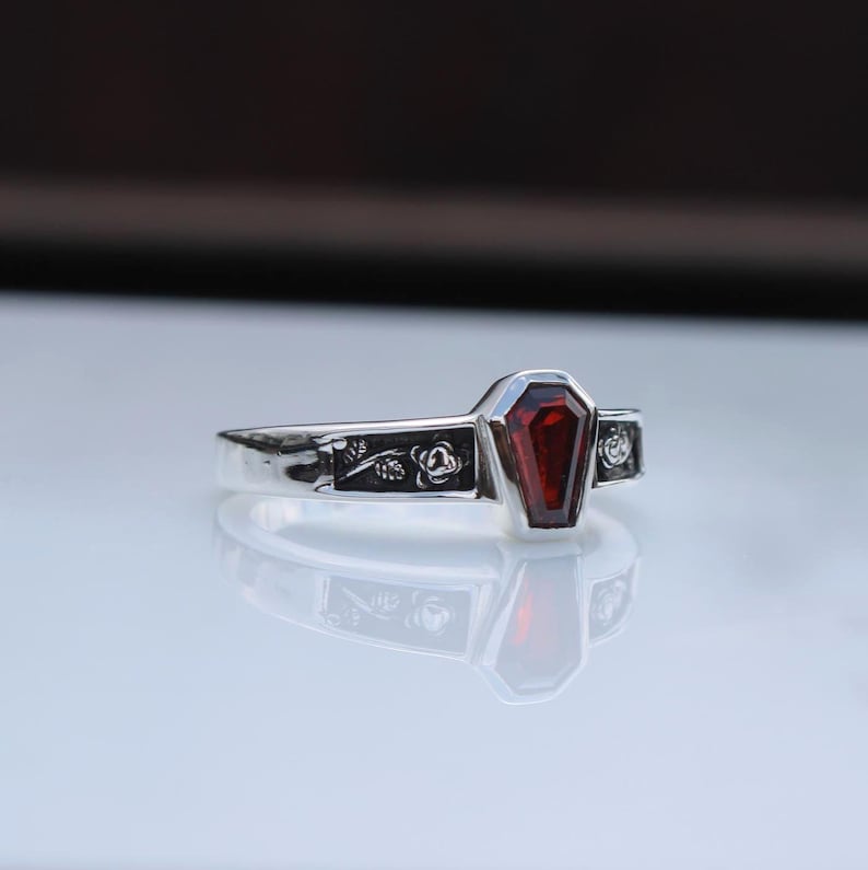 K&ouml;nnte beinhalten: Ein silberner Ring mit einem Sarg-f&ouml;rmigen, tiefroten Edelstein. Der Ring hat florale und dekorative Details am Band. Der Edelstein ist in einer silbernen L&uuml;nette gefasst. Der Ring wird auf einer reflektierenden Oberfl&auml;che pr&auml;sentiert.
