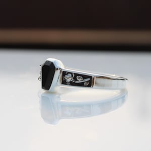 K&ouml;nnte beinhalten: Ein silberner Ring mit einem schwarzen, sargf&ouml;rmigen Stein. Der Ring hat ein dekoratives rechteckiges Feld mit floralem Design. Der Ring wird auf einer reflektierenden Oberfl&auml;che gezeigt.