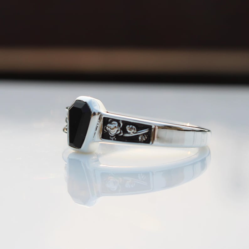 K&ouml;nnte beinhalten: Ein silberner Ring mit einem schwarzen, sargf&ouml;rmigen Stein. Der Ring hat ein dekoratives rechteckiges Feld mit floralem Design. Der Ring wird auf einer reflektierenden Oberfl&auml;che gezeigt.