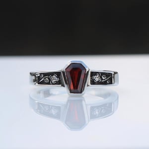 K&ouml;nnte beinhalten: Ein silberner Ring mit einem Sarg-f&ouml;rmigen, tiefroten Edelstein. Das Band ist mit floralen Gravuren verziert. Der Ring wird auf einer reflektierenden Oberfl&auml;che gezeigt, die sein Design hervorhebt.