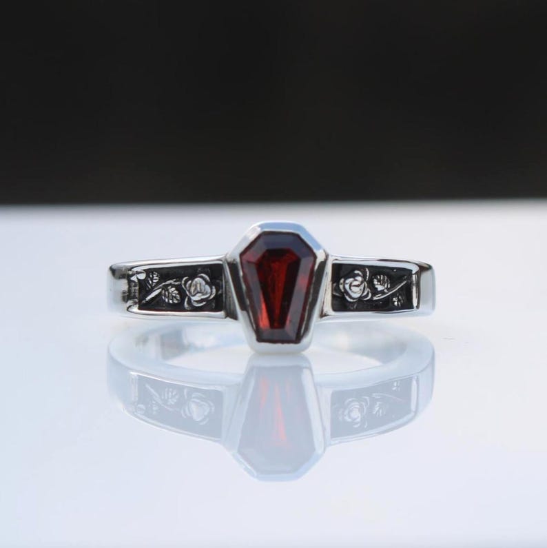 K&ouml;nnte beinhalten: Ein silberner Ring mit einem Sarg-f&ouml;rmigen, tiefroten Edelstein. Das Band ist mit floralen Gravuren verziert. Der Ring wird auf einer reflektierenden Oberfl&auml;che gezeigt, die sein Design hervorhebt.