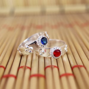 May include: Two silver rings with a blue and red gemstone. The rings are on a woven bamboo surface.
