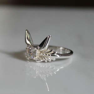 May include: A silver ring with a clear gemstone and a detailed design resembling wings.