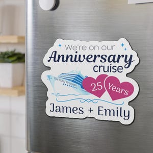 Op de afbeelding: Een witte magneet met blauwe tekst die "We're on our Anniversary cruise" zegt met een blauwe en witte cruise-scheepsafbeelding en twee roze harten die "25 Years" en "James + Emily" zeggen.