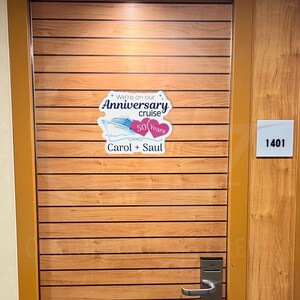 Op de afbeelding: Een houten deur met een sticker waarop staat "We're on our Anniversary cruise 50 Years Carol + Saul". De deur heeft het nummer "1401" aan de zijkant.